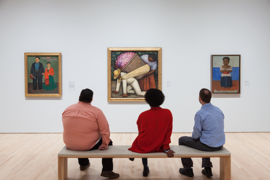 People sitting on a bench in front of Kahlo and Rivera works