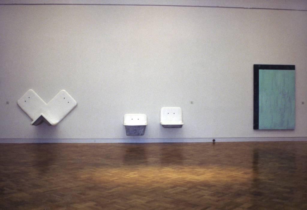 White, sink-like porcelain sculptures hang on a wall next to a large painting featuring a textured blue block and a black border around half. 